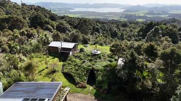 This image shows a lush, green landscape with forested hills, a small cabin, and a unique earth-sheltered home. In the distance, a tranquil lake sparkles.