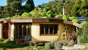 This image shows a unique eco-friendly house with a green roof covered in plants, surrounded by lush vegetation, solar panels, and garden furniture in a natural setting.