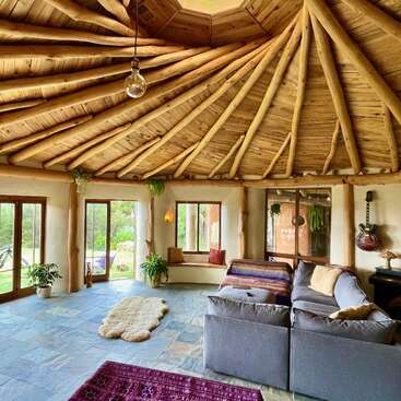 Ce salon confortable et rustique est doté d'un superbe plafond en bois, d'une lumière naturelle, de plantes luxuriantes, d'un canapé sectionnel gris, d'une guitare sur le mur et d'une vue sereine sur l'extérieur.