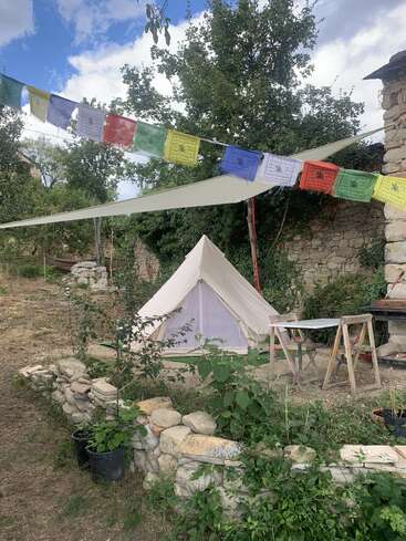 Un campement confortable est composé d'une tente en toile blanche, de drapeaux de prière colorés, de murs en pierre rustiques, de plantes vertes, d'une table de fortune et d'arbres, le tout sous un ciel partiellement nuageux.