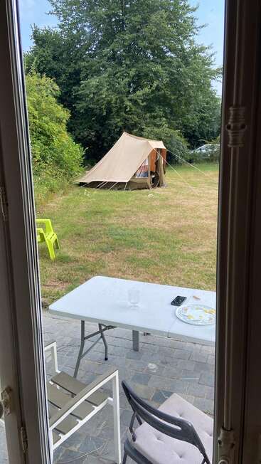 Ein Blick von drinnen zeigt eine Terrasse mit einem weißen Tisch, Stühlen, Gläsern und einem Teller. Draußen steht ein beiges Zelt auf dem Rasen in der Nähe von grünen Bäumen und gelben Stühlen.