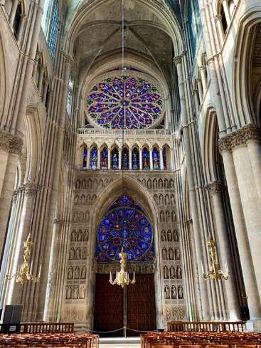 Dieses Bild zeigt das Innere einer prächtigen Kathedrale mit hoch aufragenden Gewölbedecken, kunstvollen Säulen, eleganten Kronleuchtern und atemberaubenden Buntglas-Rosettenfenstern über einem Bogenportal.