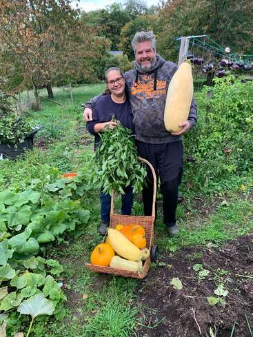 Ein lächelndes Paar steht stolz in einem üppigen Garten, hält frisch geerntetes Gemüse und Kräuter in den Händen, und zu ihren Füßen steht ein Korb mit Kürbissen und Zucchini.