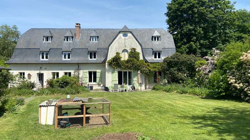 Ein charmantes Landhaus mit Schieferdach, grünen Fensterläden und einem üppigen Garten. Im Vordergrund ein Hühnerstall, umgeben von sattem Grün, Bäumen und blühenden Blumen an einem sonnigen Tag.