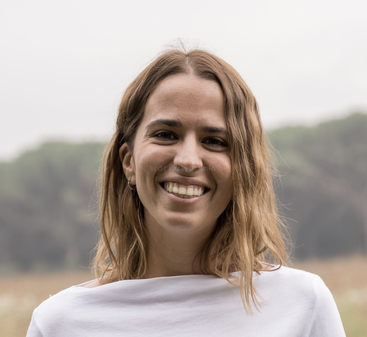 A young woman with shoulder-length light brown hair smiles warmly outdoors, wearing a white top. The background is blurry with trees and a soft, natural setting.