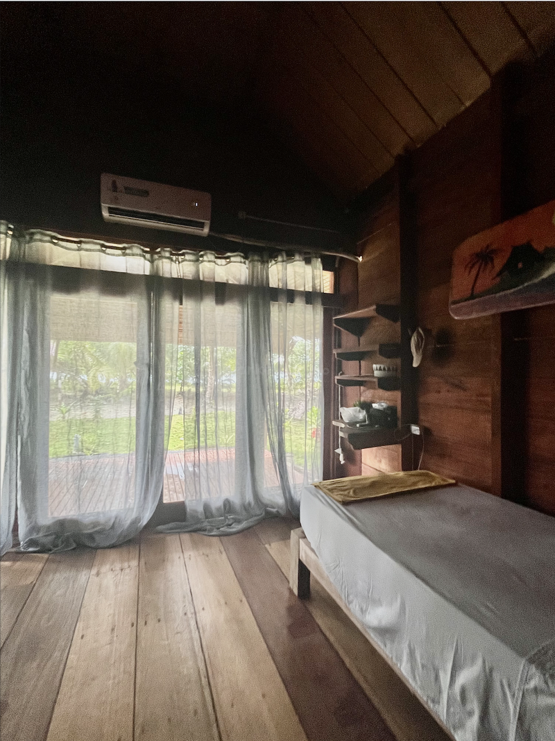 Uma aconchegante sala de madeira com uma mesa de massagem, cortinas transparentes que deixam a luz natural entrar, prateleiras de madeira e uma vista para a vegetação lá fora, evocando relaxamento e tranquilidade.