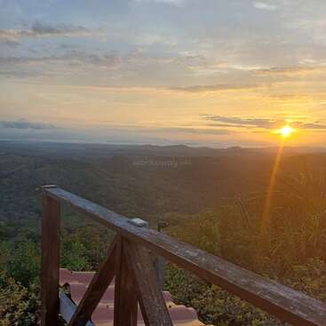 A imagem captura um pôr do sol de tirar o fôlego sobre colinas onduladas. A luz do sol ilumina as nuvens e a paisagem. Uma grade de madeira e um telhado de telhas aparecem em primeiro plano. Cenário tranquilo.