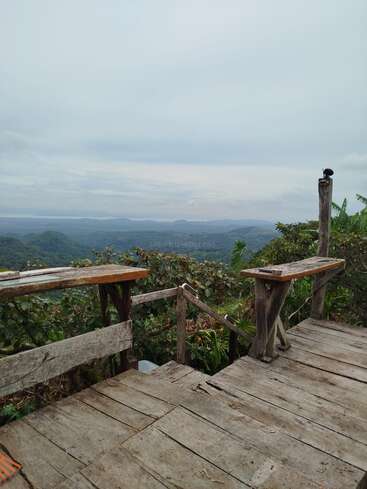 Eine rustikale Holzterrasse blickt auf üppig grüne Hügel unter einem wolkenverhangenen Himmel. Einfache Bänke laden zur stillen Einkehr ein, umgeben von der Schönheit der Natur und fernen, sanften Landschaften.