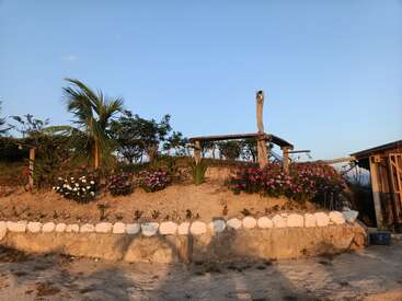 Ein sandiger Gartenhügel mit weißen Steinen grenzt an bunte Blumen und tropische Pflanzen. Obenauf steht eine hölzerne Pergola, die unter dem klaren Himmel von warmem, goldenem Sonnenlicht berührt wird.