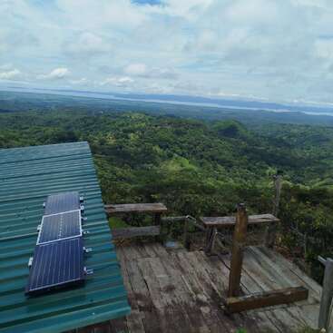 A wooden deck overlooks a lush green valley with distant water. A corrugated metal roof with solar panels is visible under a partly cloudy sky.