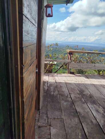 Um deck de madeira rústica se abre para uma vista deslumbrante de colinas verdes e céu azul, vista por uma porta ligeiramente aberta. As nuvens flutuam suavemente acima de você.