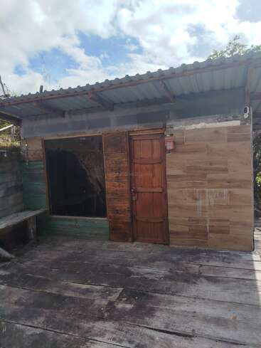 Uma pequena estrutura rústica com materiais de madeira e incompatíveis, uma porta simples e um telhado de metal corrugado. A área tem um piso de madeira, um banco e um céu nublado acima.