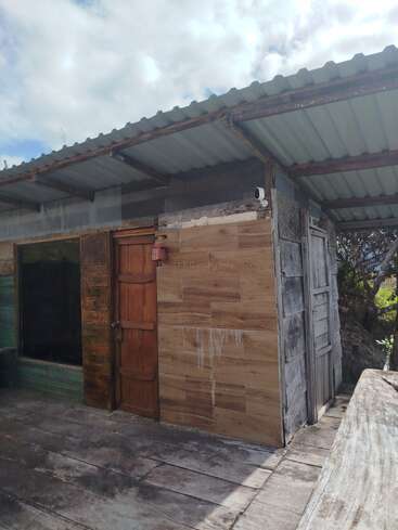 Esta imagem mostra um pequeno edifício rústico com telhado de zinco, portas de madeira, parede parcialmente revestida de azulejos, câmera de segurança e deck de madeira desgastada sob um céu nublado.