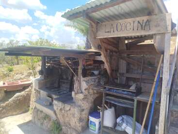 Cette image montre une cuisine extérieure rustique intitulée « La Cocina », avec des plans de travail en pierre, un abri en bois, un barbecue en métal, divers ustensiles et un paysage champêtre en arrière-plan.