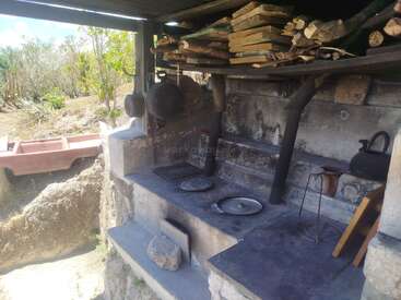 Cette image montre une cuisine rustique en plein air avec un poêle à bois, des casseroles suspendues, un tas de bois de chauffage au-dessus, des bouilloires en métal, le tout sur fond de paysage naturel et champêtre baigné de soleil.