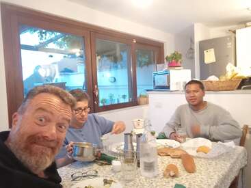 Das Bild zeigt drei Personen, die an einem Tisch sitzen: ein Mann im Vordergrund, eine Frau hinter ihm und ein Mann auf der rechten Seite, die alle mit dem Essen beschäftigt sind.