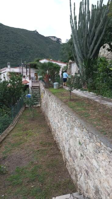 Das Bild zeigt einen ruhigen, von Steinmauern gesäumten Weg in einer bergigen Gegend, auf dem eine Person von einem Haus weggeht, umgeben von üppigem Grün und einem majestätischen Berg im Hintergrund.