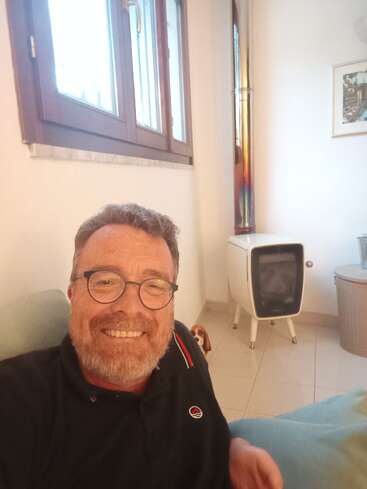 The image depicts a man sitting in a room with white walls, a window, and a small dog, accompanied by a white stove and a framed picture on the wall behind him.