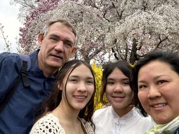 A family of four smiles for a selfie outdoors, surrounded by blooming cherry blossoms and colorful spring flowers, enjoying a beautiful, cheerful day together in nature.