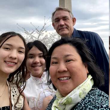 Uma família de quatro pessoas sorri para uma foto ao ar livre. As flores de cerejeira florescem atrás deles. O Monumento a Washington surge ao fundo em um dia nublado.