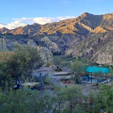 De majestueuses montagnes baignées de soleil s'élèvent derrière une vallée. Des arbres et des arbustes parsèment le premier plan, tandis que des pavillons colorés et des bâtiments pittoresques sont nichés dans le paysage. Scène paisible.