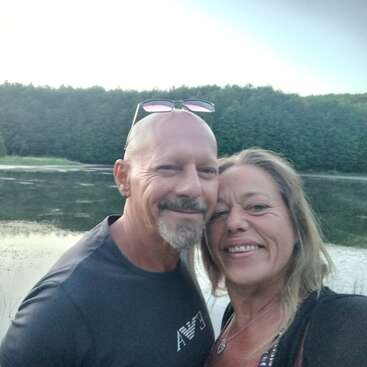 Um homem e uma mulher sorridentes tiram uma selfie juntos ao ar livre perto de um lago sereno, cercados por árvores verdes exuberantes, desfrutando de um momento tranquilo e feliz juntos.