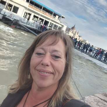 The image shows a woman taking a selfie in front of a large boat on a river, with a bridge and city skyline in the background.