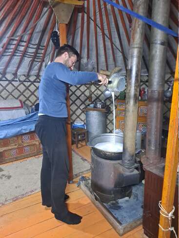 Ein Mann in einer traditionellen Jurte gießt Flüssigkeit aus einem Krug in einen Topf auf dem Herd. Der gemütliche Innenraum ist mit Holzmöbeln und buntem Dekor ausgestattet.