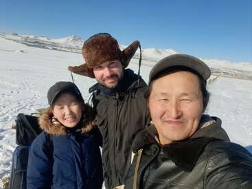 Drei Menschen lächeln im Freien in einer verschneiten Landschaft unter einem klaren blauen Himmel. Sie tragen warme Winterkleidung, darunter Mützen und Mäntel, und im Hintergrund sind die Berge zu sehen.