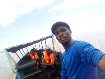 Das Bild zeigt einen Mann, der ein Selfie auf einem Boot macht, mit drei Personen in orangefarbenen Schwimmwesten im Hintergrund, vor einer ruhigen Wasseroberfläche.