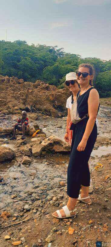 Deux femmes portant des lunettes de soleil sourient à l'appareil photo, debout sur un terrain rocheux avec de la verdure derrière. Un enfant est assis à l'arrière-plan, près de rochers et d'eau.