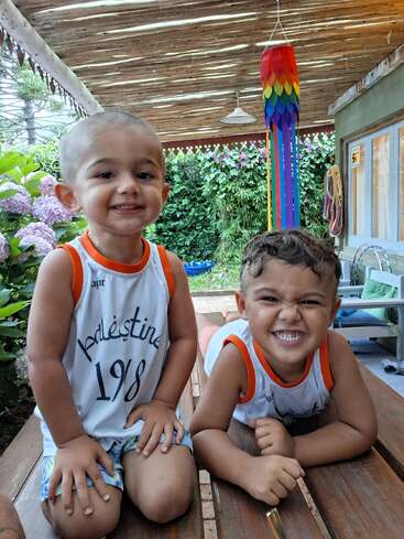Zwei kleine Kinder grinsen fröhlich auf einem Holztisch unter einer überdachten Terrasse und tragen die gleichen Shirts. Bunte Deko und viel Grün sorgen für eine fröhliche Outdoor-Atmosphäre.