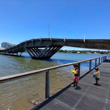 Two children stand on a walkway beside a unique, wavy concrete bridge over water. The sky is clear and blue, suggesting a sunny, pleasant day.