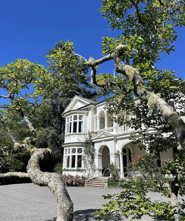 Une belle maison blanche de style victorien est baignée de soleil, entourée de verdure luxuriante, d'arbres aux branches moussues et de fleurs épanouies dans un ciel bleu limpide.
