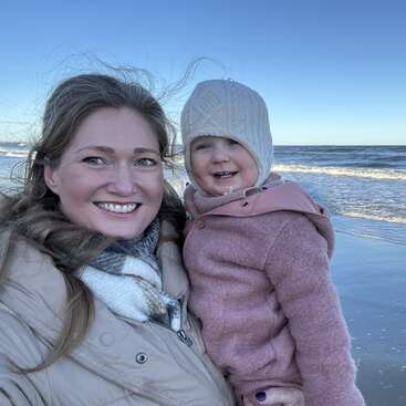 A smiling woman and a young child bundled in warm jackets pose together on a beach, with waves and a clear blue sky in the background.