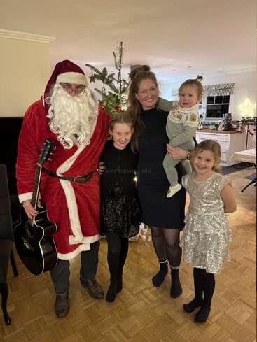 A family poses indoors with someone dressed as Santa Claus holding a guitar. Behind them is a decorated Christmas tree. Everyone appears happy, celebrating together.