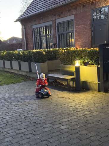 A small child in a red coat rides a toy car on a cobblestone path outside a house, illuminated by warm garden lights at dusk.