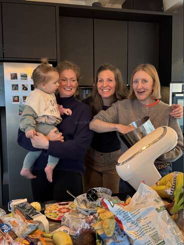 Three women and a toddler happily gather in a kitchen, surrounded by snacks and ingredients, as one woman operates a stand mixer, preparing food together.