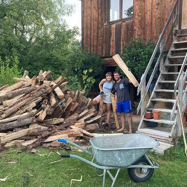 L'image représente un couple debout devant un grand tas de bois de chauffage, avec une brouette au premier plan et une maison en bois à l'arrière-plan.