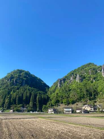 Essa imagem mostra uma linda paisagem rural com colinas verdes, algumas casinhas, campos em primeiro plano e um céu azul claro e brilhante acima.