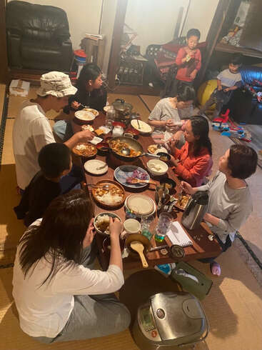 Uma família se reúne em torno de uma mesa baixa, saboreando uma refeição em conjunto numa sala aconchegante, com as crianças brincando por perto, criando juntos um ambiente acolhedor e animado.