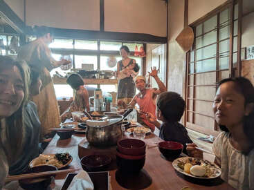 Uma família se reúne em torno de uma mesa baixa tradicional, saboreando uma refeição juntos em uma aconchegante sala de estilo japonês, banhada pela luz do sol. Risos e calor humano preenchem o ambiente.