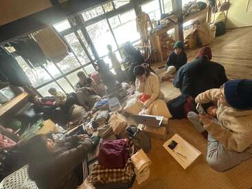 Um grupo de pessoas se reúne dentro de casa, cercado por roupas e pertences, relaxando e conversando perto de grandes janelas num dia de neve, criando um ambiente aconchegante e de convívio.