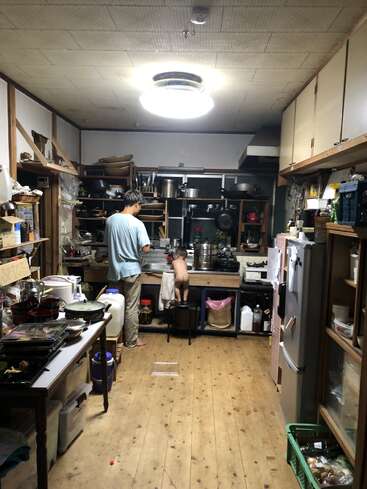 Um homem e uma criança pequena, em cima de um banquinho, estão ocupados na pia de uma cozinha desarrumada, aconchegante e bem usada, com piso de madeira.