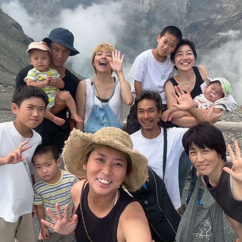 Um grupo alegre de pessoas, incluindo adultos e crianças, posa ao ar livre em frente a um cenário montanhoso envolto em névoa, sorrindo e acenando para a câmera.