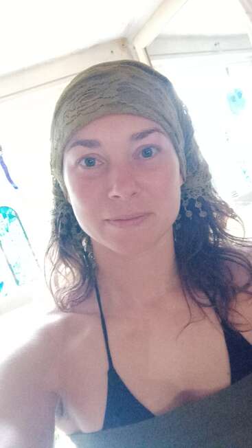 Eine Frau mit gewelltem Haar trägt ein grünes Kopftuch und ein schwarzes Oberteil. Sie steht in einem Innenraum, in dem helles Tageslicht und Buntglas im Hintergrund zu sehen sind.
