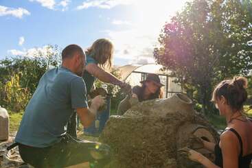 Cuatro personas trabajan juntas al aire libre, dando forma a una gran estructura de arcilla y paja bajo un sol radiante, rodeadas de árboles y naturaleza, haciendo gala de su espíritu de equipo y de prácticas de construcción sostenible.