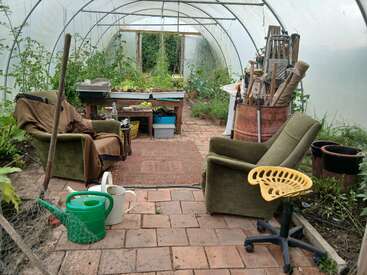 Un acogedor invernadero con dos sillones, herramientas de jardinería, regaderas y plantas frondosas. El camino de ladrillos y las alfombras aportan calidez, creando un espacio de jardinería relajante y práctico.