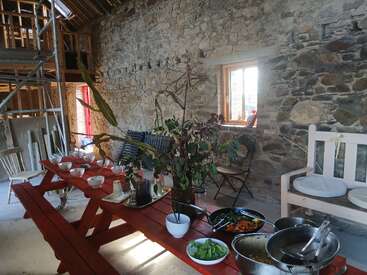 Una habitación rústica con paredes de piedra en la que se ve una mesa de picnic roja preparada para comer, con cuencos, ensalada y un centro de mesa con plantas, todo ello bajo un techo de madera a medio construir.