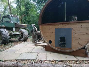 Un tractor verde con neumáticos grandes está aparcado junto a una sauna redonda de madera en una zona boscosa, sobre baldosas de piedra rodeadas de árboles y vegetación.